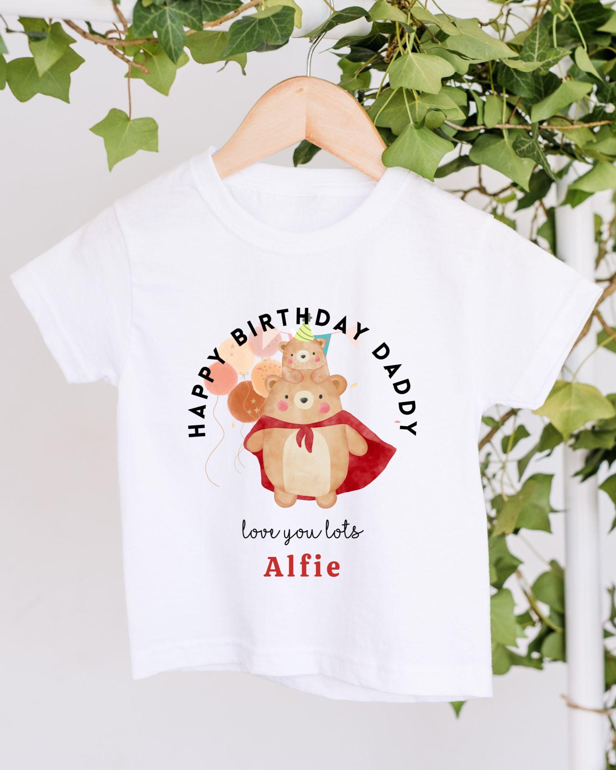 A white t-shirt with a graphic design featuring a bear wearing a red cape and a birthday hat, along with the text "Happy Birthday Daddy" and "love you lots Alfie".