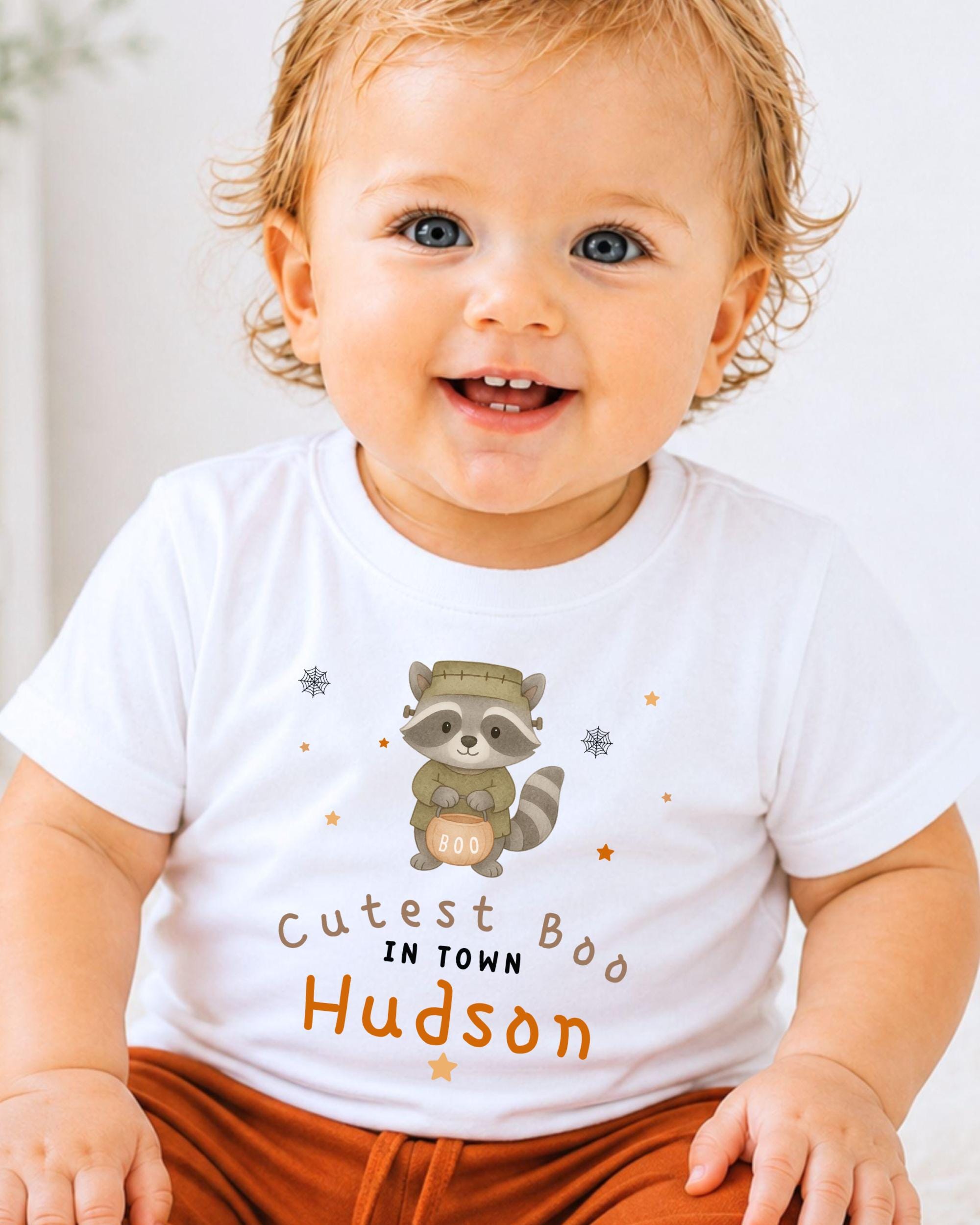 A smiling baby wearing a white t-shirt with a raccoon graphic and text that says "Cute Boo in Town Hudson".