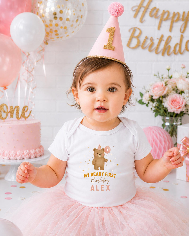 A young girl wearing a birthday hat and a white t-shirt with a bear on it, sitting in front of a birthday cake and balloons.