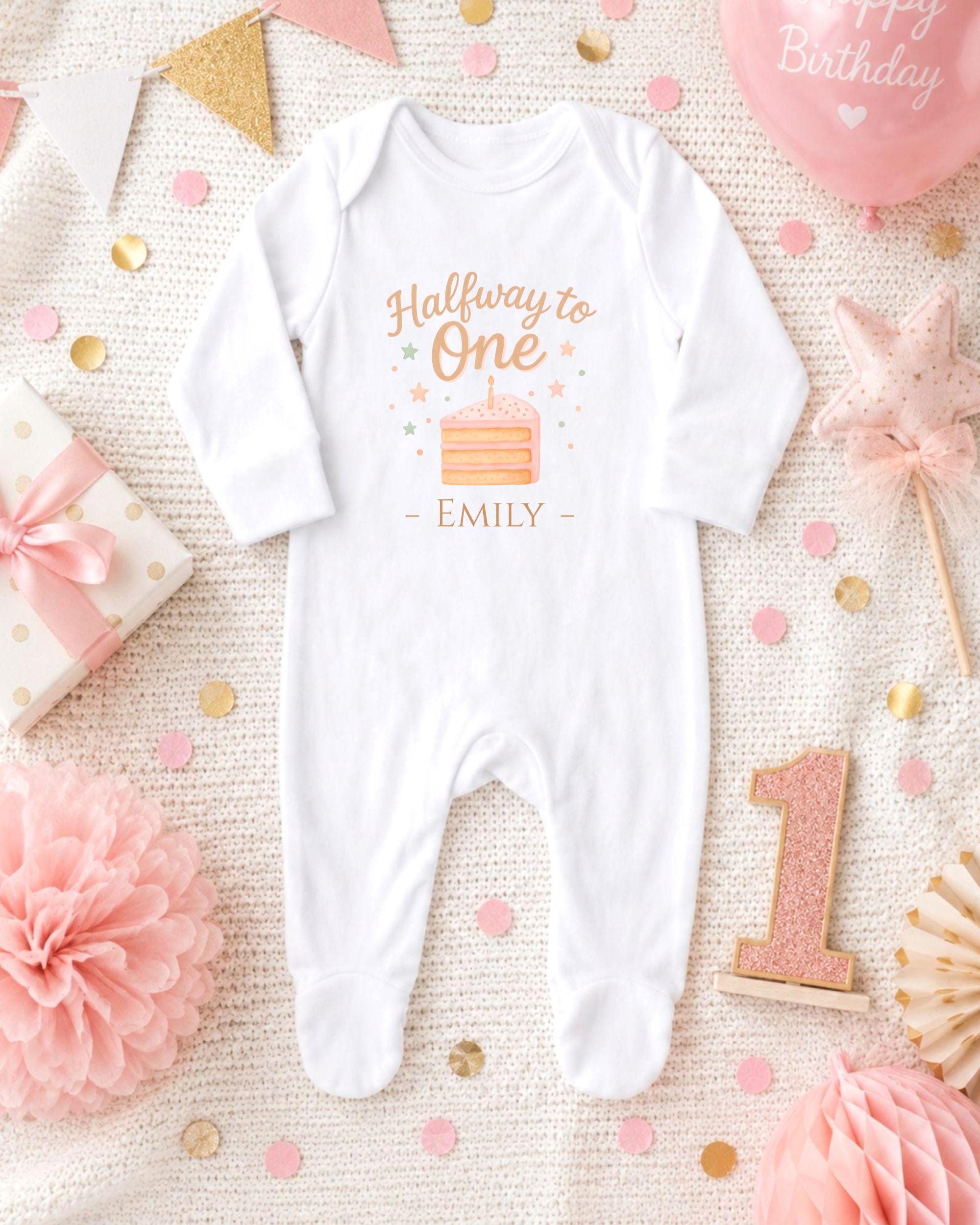 A white onesie with a birthday message printed on it, surrounded by pink and gold decorations, confetti, and a pink balloon.