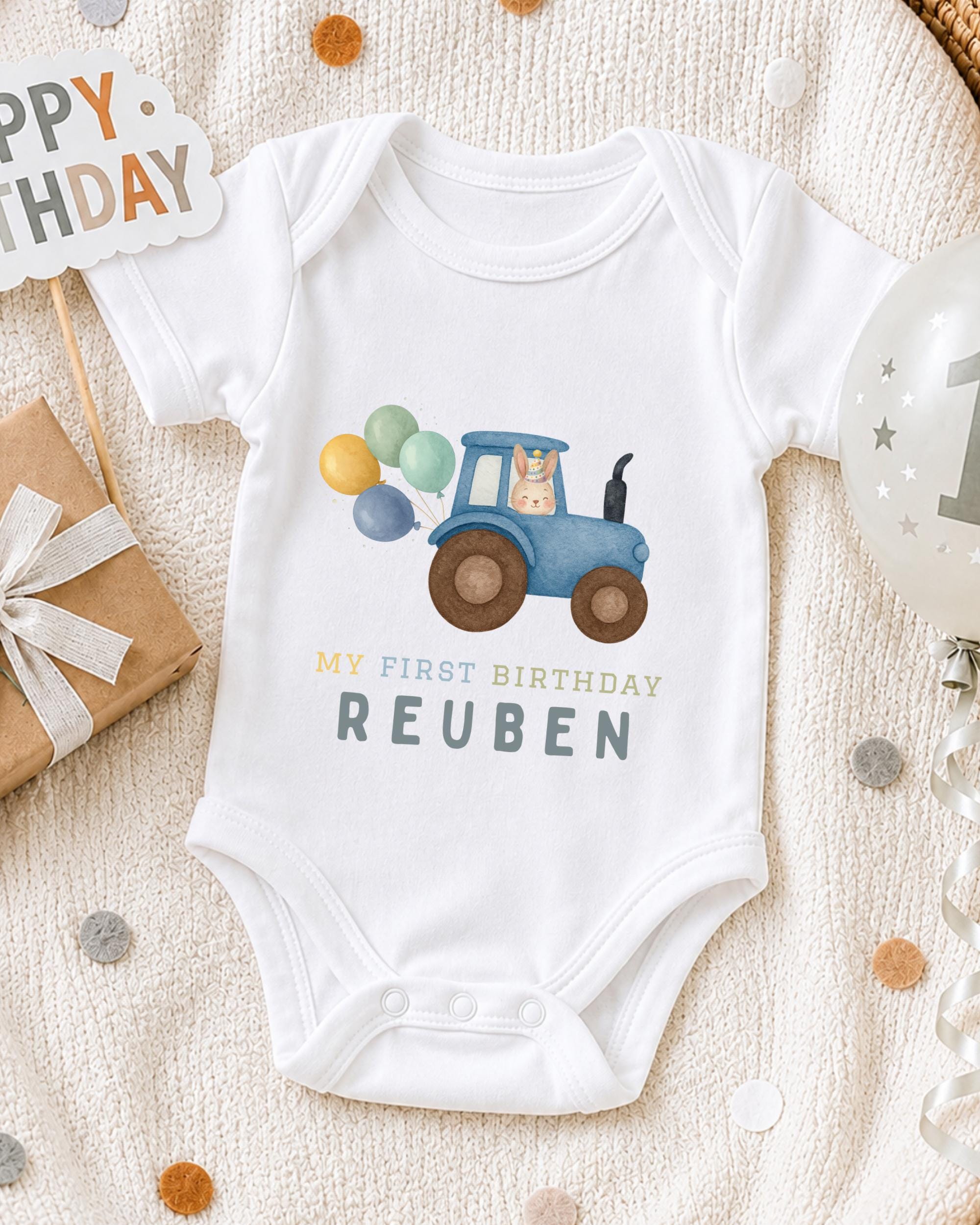 A white onesie with a blue tractor and balloons on it, along with a "Happy Birthday Reuben" text, is displayed on a bed with confetti.