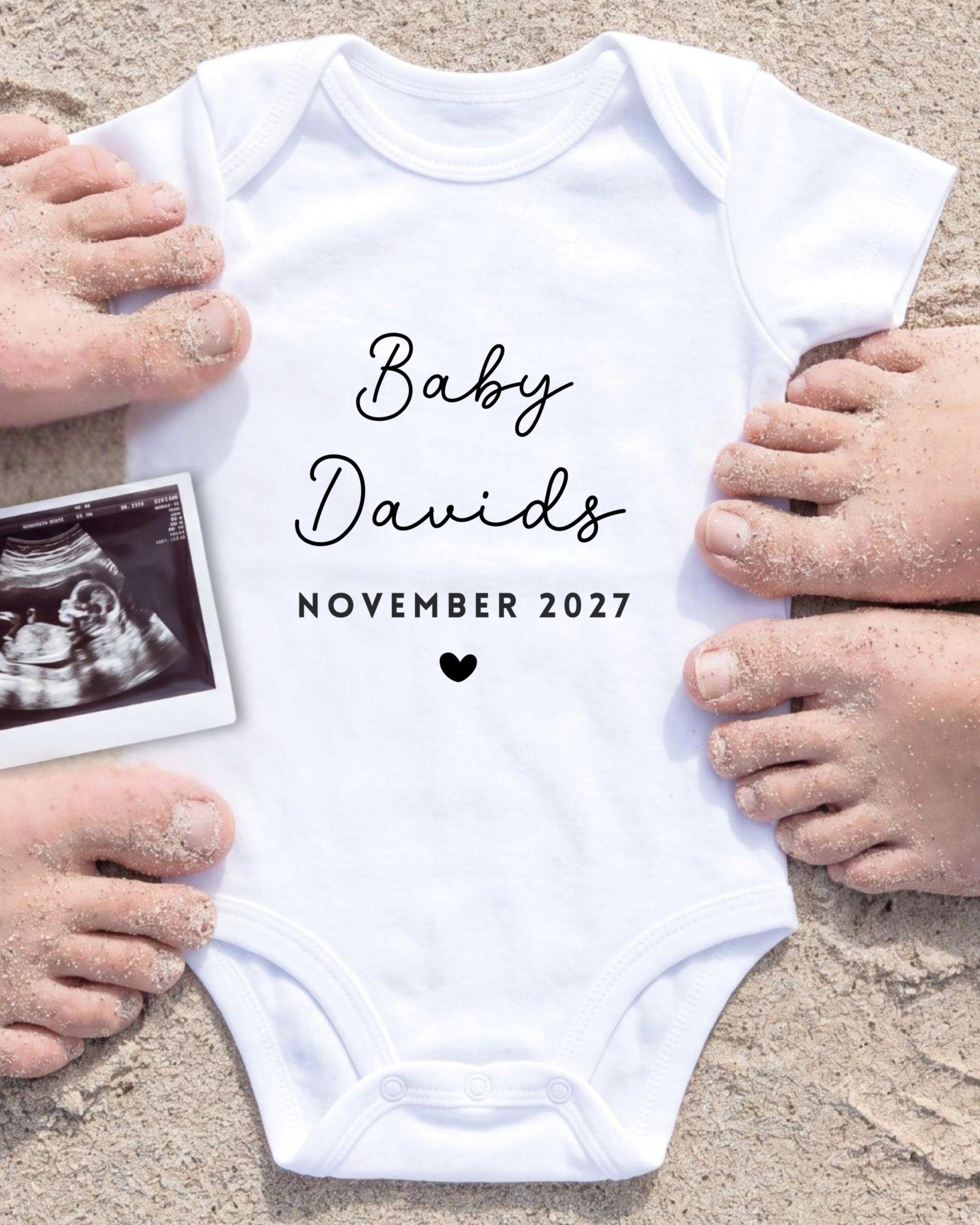 A white baby onesie with the text "Baby Davis" and "November 2027" printed on it, surrounded by sand and a black and white photograph of a baby's belly.