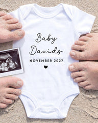 A white baby onesie with the text "Baby Davis" and "November 2027" printed on it, surrounded by sand and a black and white photograph of a baby's belly.