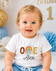 A smiling baby boy wearing a white t-shirt with a rainbow graphic and the name "Alex" printed on it, sitting in front of a birthday party backdrop.