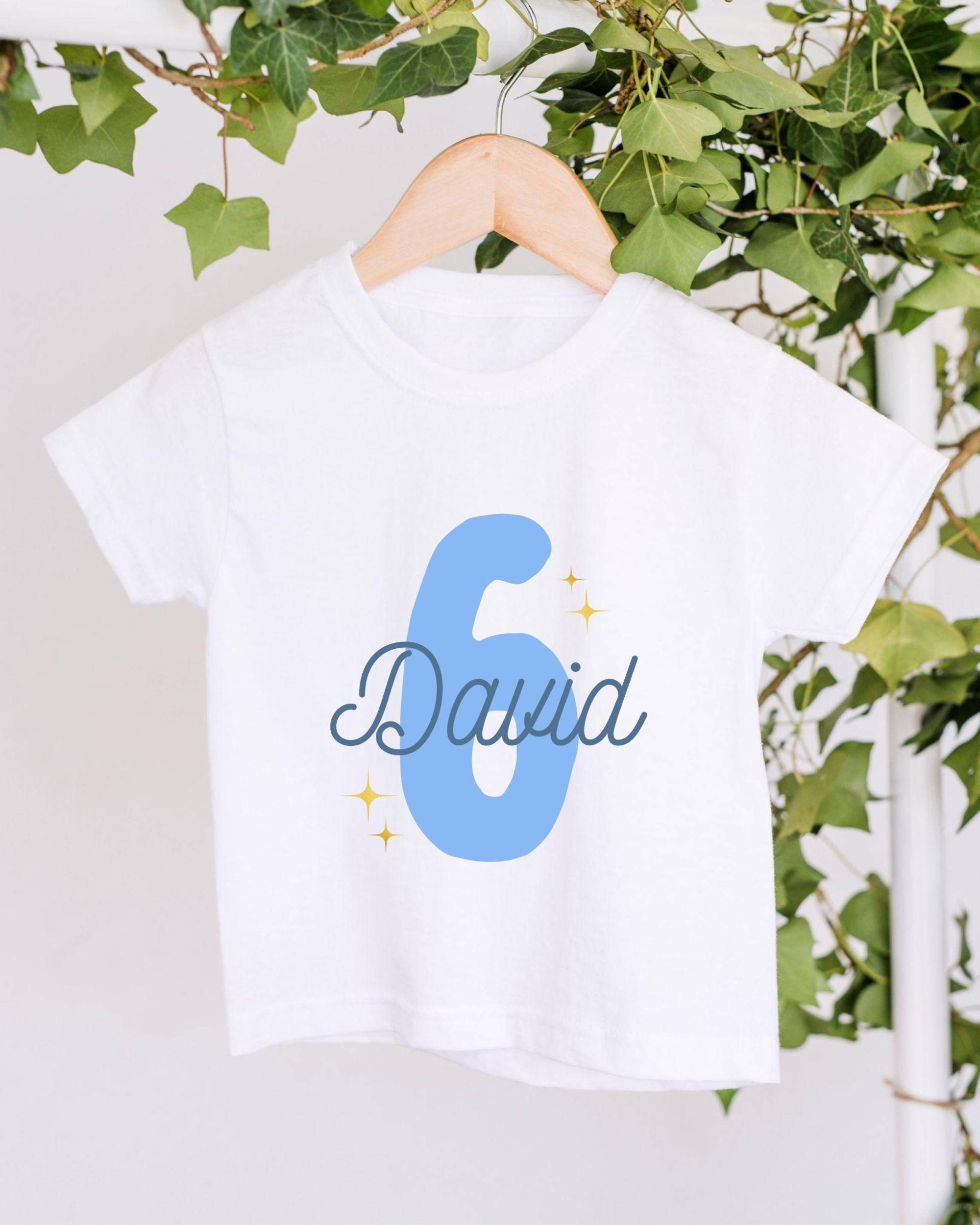 A white t-shirt with the number 6 and the name "David" printed on it, hanging on a wooden hanger.