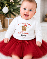 A smiling baby girl wearing a white sweater with a teddy bear graphic and text, sitting on a red tulle skirt in front of a Christmas tree.