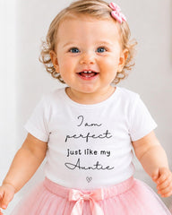 A smiling baby girl wearing a white t-shirt with text and a pink bow in her hair.