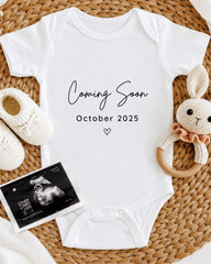 A white baby onesie with the text "Coming Soon October 2025" printed on it, along with a crocheted bunny toy and a black and white photograph of an infant's head.