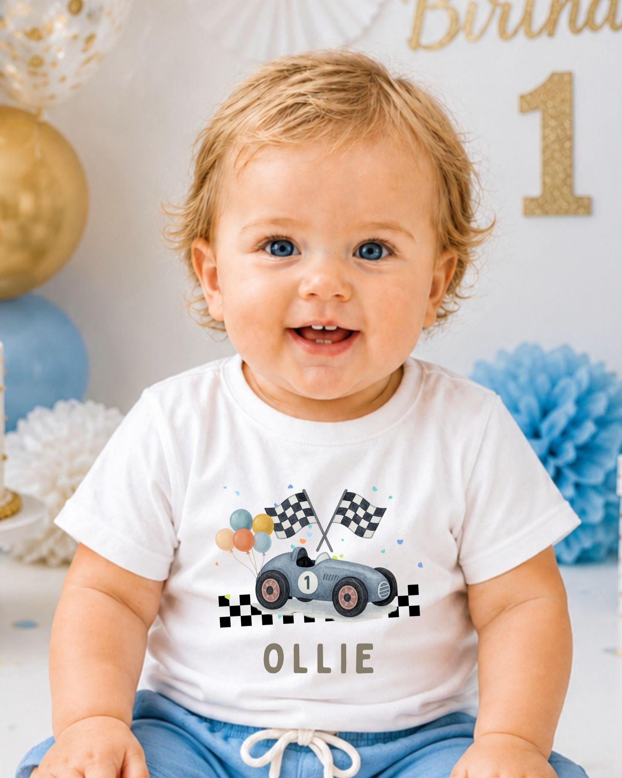 A smiling baby boy wearing a white t-shirt with a race car graphic and the name "Ollie" printed on it, sitting in front of a birthday party backdrop.