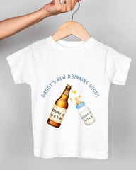 A white t-shirt with a graphic design depicting a beer bottle and a baby bottle, with the text "DADDY'S NEW DRINKING BUDDY" printed on the shirt.