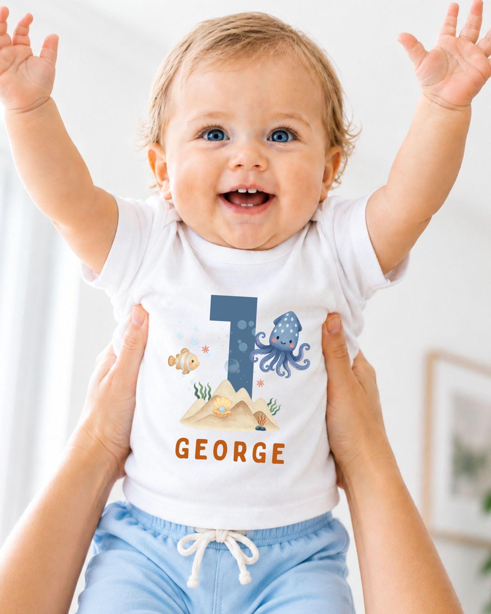 A smiling baby wearing a white shirt with a blue number 1 and an underwater-themed design, being held up by someone's arms.