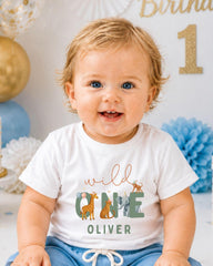 A smiling baby boy wearing a white t-shirt with a graphic design of wild animals and the text "wild one Oliver".