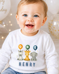 A smiling baby boy wearing a white shirt with a cartoon image of three animals and the name "HENRY" printed on it.