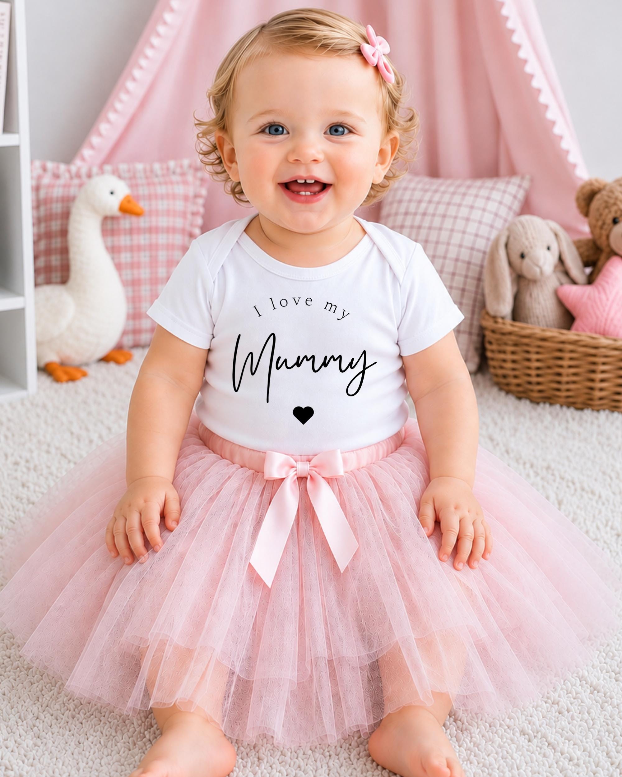A smiling baby girl wearing a white t-shirt with the text "I love my Mummy" and a pink tutu skirt, sitting on a white carpet surrounded by stuffed animals.