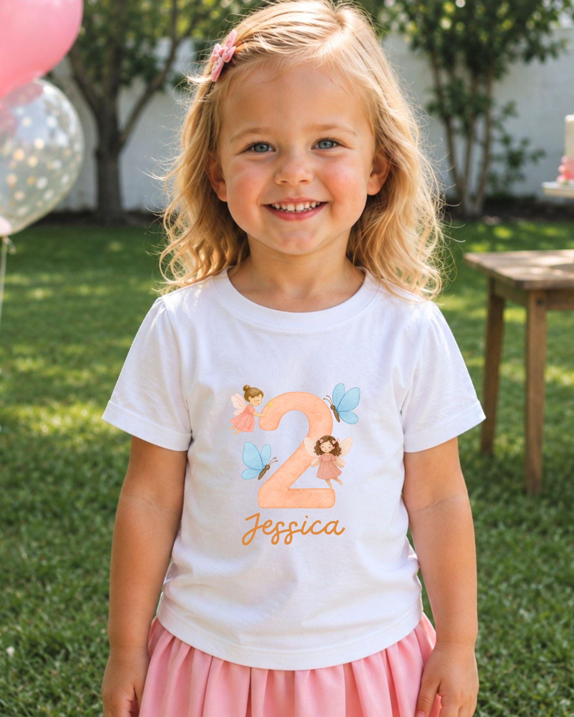 A young girl with blonde hair is smiling and wearing a white t-shirt with a personalized number 2 and fairy design printed on it.
