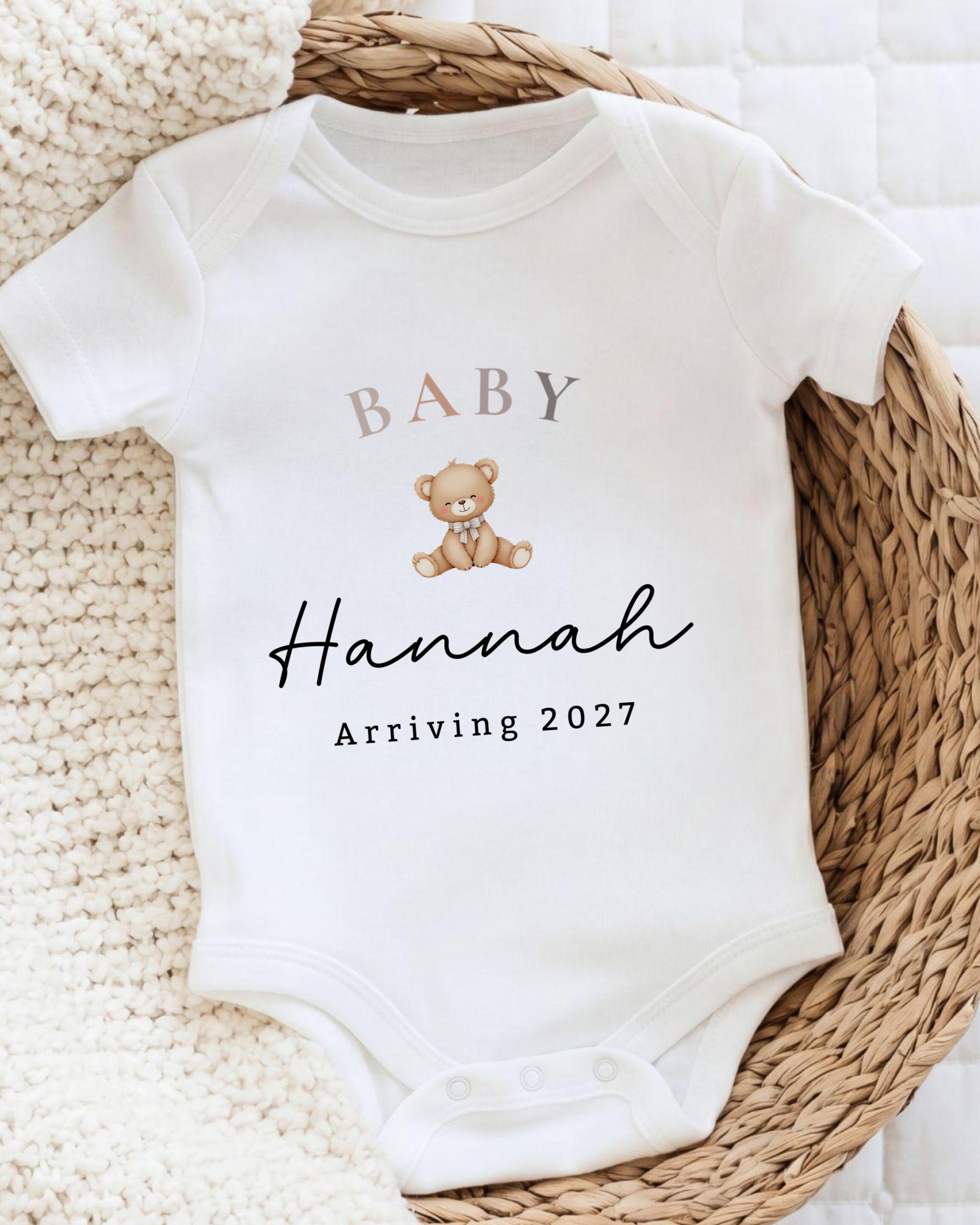 A white baby onesie with the text "BABY Hannah Arriving 2027" printed on it, along with a teddy bear graphic.