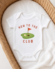 A white onesie with the text "NEW TO THE CLUB" printed on it, along with a golf hole graphic, is displayed on a white blanket.