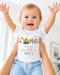 A smiling baby wearing a white shirt with a construction vehicle graphic and the text "One busy builder Matt" is being held up by someone's arms.