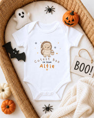 A white onesie with a cartoon character and text, surrounded by Halloween decorations such as a ghost, a pumpkin, a bat, and a sign that says "BOO!".