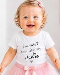 A smiling baby girl wearing a white t-shirt with text and a pink bow in her hair.