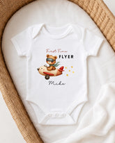 A white onesie with a cartoon image of a bear wearing a pilot's hat and flying an airplane, along with the text "First Time Flyer" and "Miki".