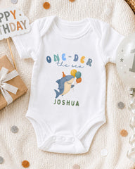 A white onesie with a shark design and the text "One-der the sea Joshua" printed on it, along with a birthday sign and balloons, suggesting a celebration for a baby's first birthday.