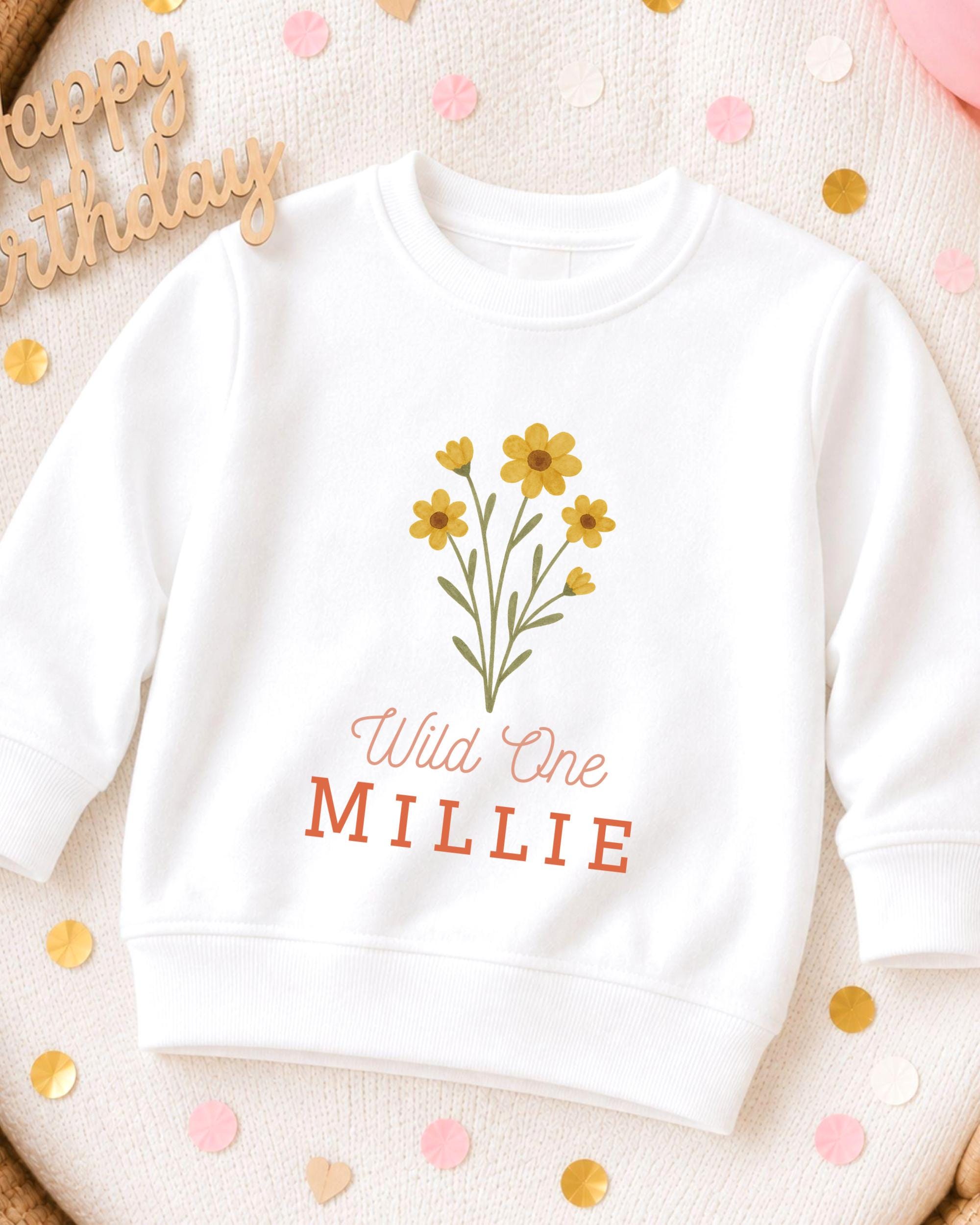 A white sweatshirt with a yellow flower design and the text "Wild One MILLIE" printed on it, along with a "Happy Birthday" greeting in the background.