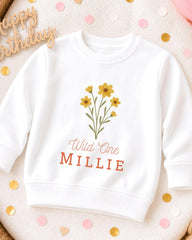 A white sweatshirt with a yellow flower design and the text "Wild One MILLIE" printed on it, along with a "Happy Birthday" greeting in the background.