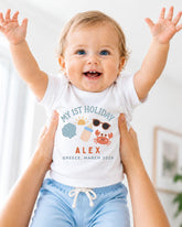 A smiling baby wearing a white t-shirt with a personalized design and text, being held up by someone's arms.