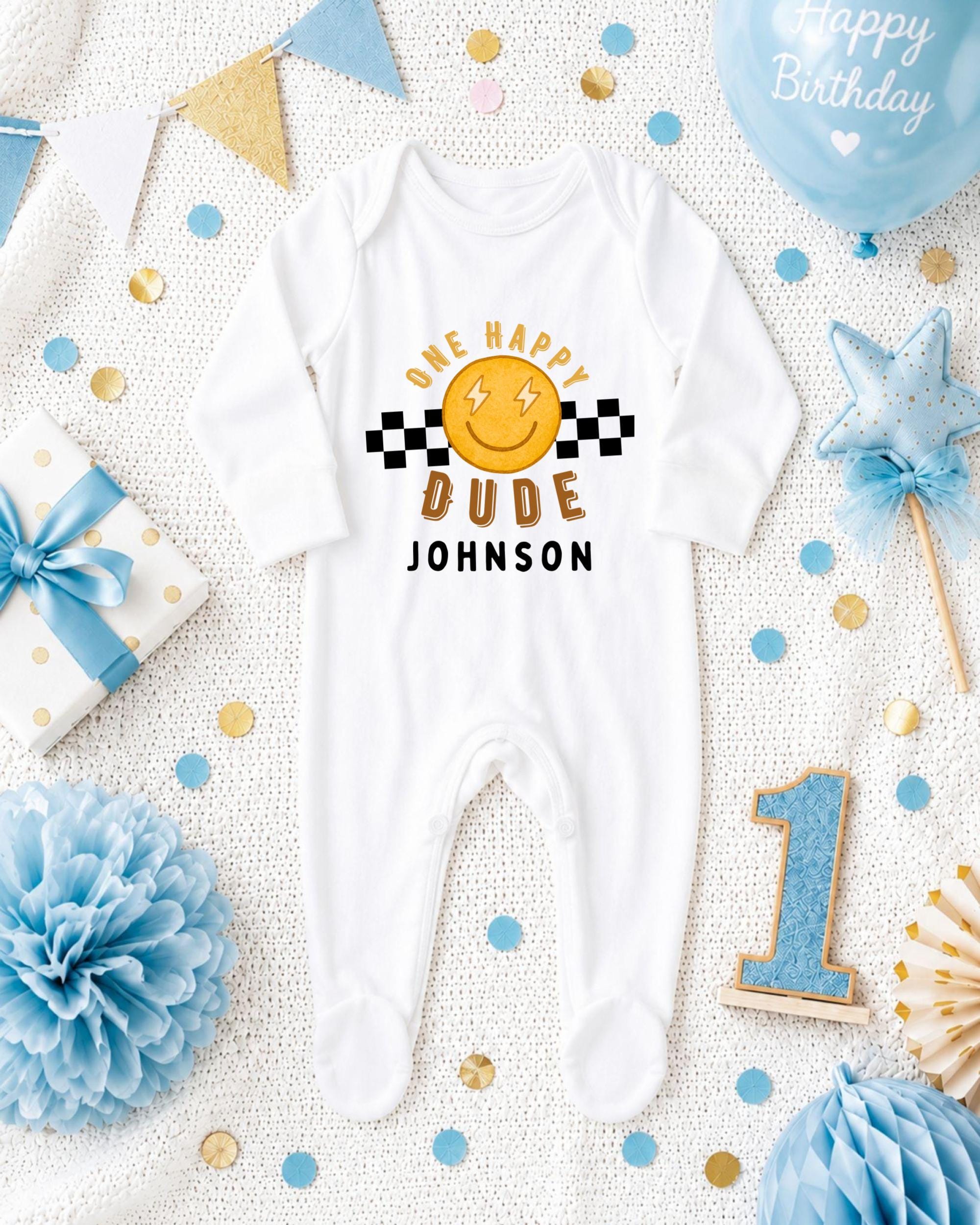 A white onesie with a smiley face and the text "One happy dude Johnson" printed on it, surrounded by birthday decorations and a blue balloon.