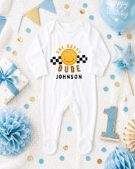 A white onesie with a smiley face and the text "One happy dude Johnson" printed on it, surrounded by birthday decorations and a blue balloon.