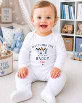 A smiling baby boy wearing a white onesie with the text "Watching the race with my daddy" sits on the floor in a nursery setting.