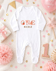 A white onesie with the name "Nicole" printed on it, surrounded by pink and gold decorations, including a birthday banner, confetti, and a star-shaped ornament.