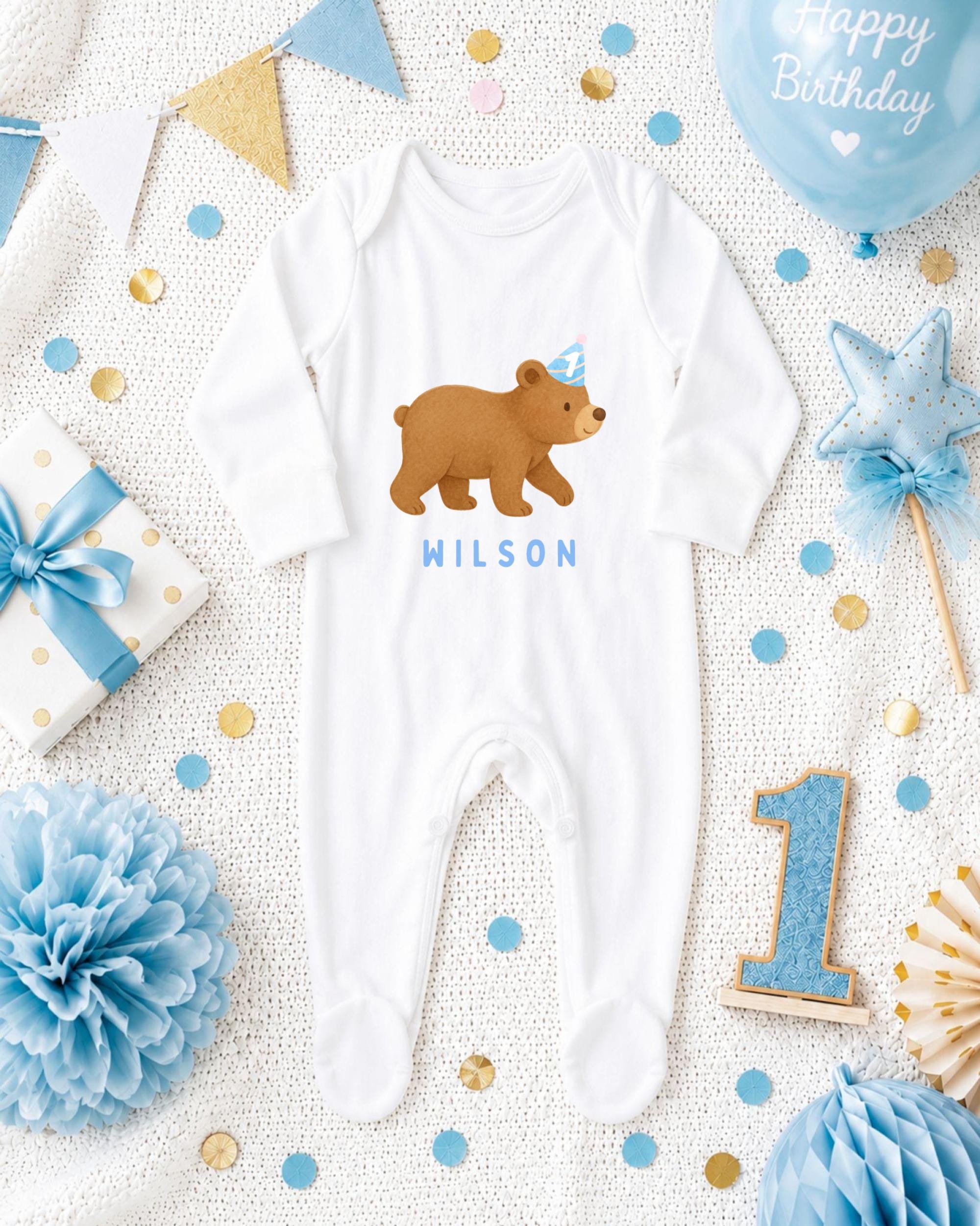 A baby's onesie with a bear design, surrounded by birthday decorations such as a blue balloon, blue and gold confetti, and a blue ribbon.