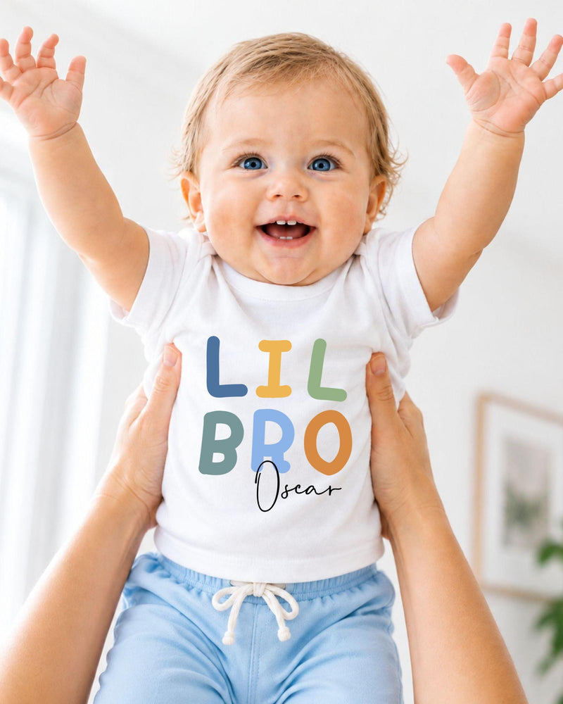 A smiling baby wearing a white t-shirt with the text "LIL BRO Oscar" is being held up by someone's arms.