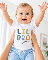 A smiling baby wearing a white t-shirt with the text "LIL BRO Oscar" is being held up by someone's arms.
