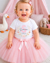 A smiling baby girl wearing a white t-shirt with a pink floral design and a pink tutu skirt, sitting on a pink carpet surrounded by stuffed animals.