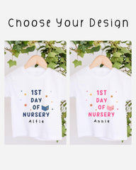 two white t-shirts hanging on a wooden hanger, each with a different design and text. The t-shirts are labeled "1st Day of Nursery" and "1st Day of Nursery" with the names "Alfie" and "Annie" printed on them. The t-shirts are set against a backdrop of greenery, creating a pleasant and natural atmosphere.