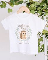 A white t-shirt with a hedgehog graphic and the text "Halfway to One - Izzy" printed on it, hanging on a wooden hanger.
