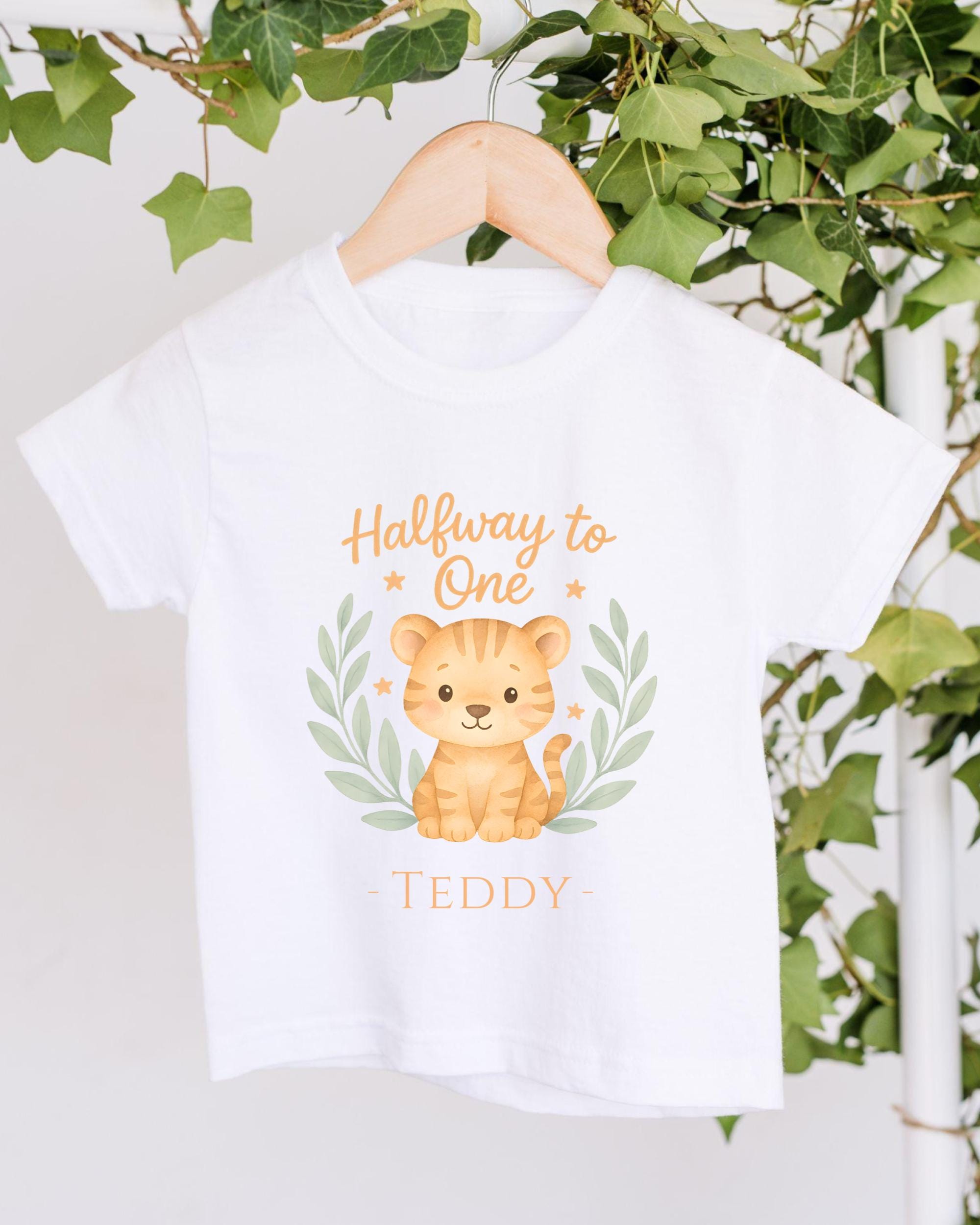 A white t-shirt with a cartoon tiger graphic and the text "Halfway to One" printed on it, hanging on a wooden hanger.