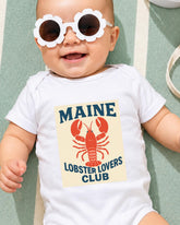 A baby wearing a white onesie with a lobster graphic and sunglasses, lying on a green surface.