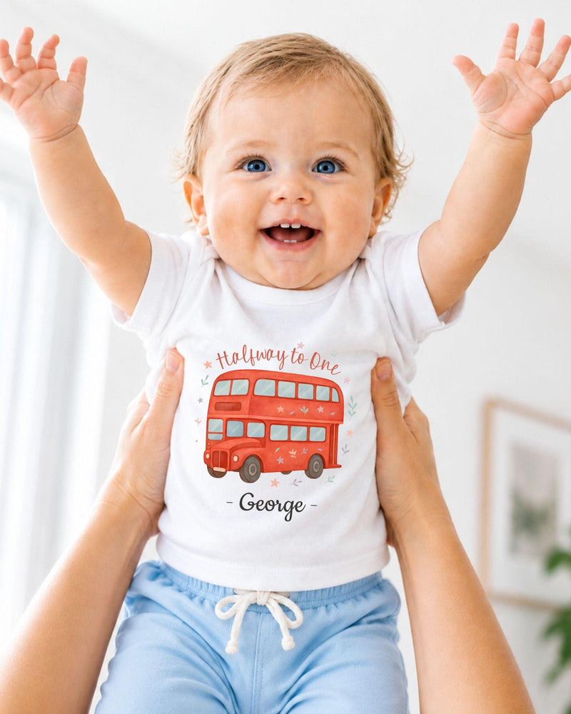A smiling baby wearing a white t-shirt with a red double-decker bus design and blue shorts, with their arms raised in a joyful gesture.
