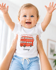 A smiling baby wearing a white t-shirt with a red double-decker bus design and blue shorts, with their arms raised in a joyful gesture.