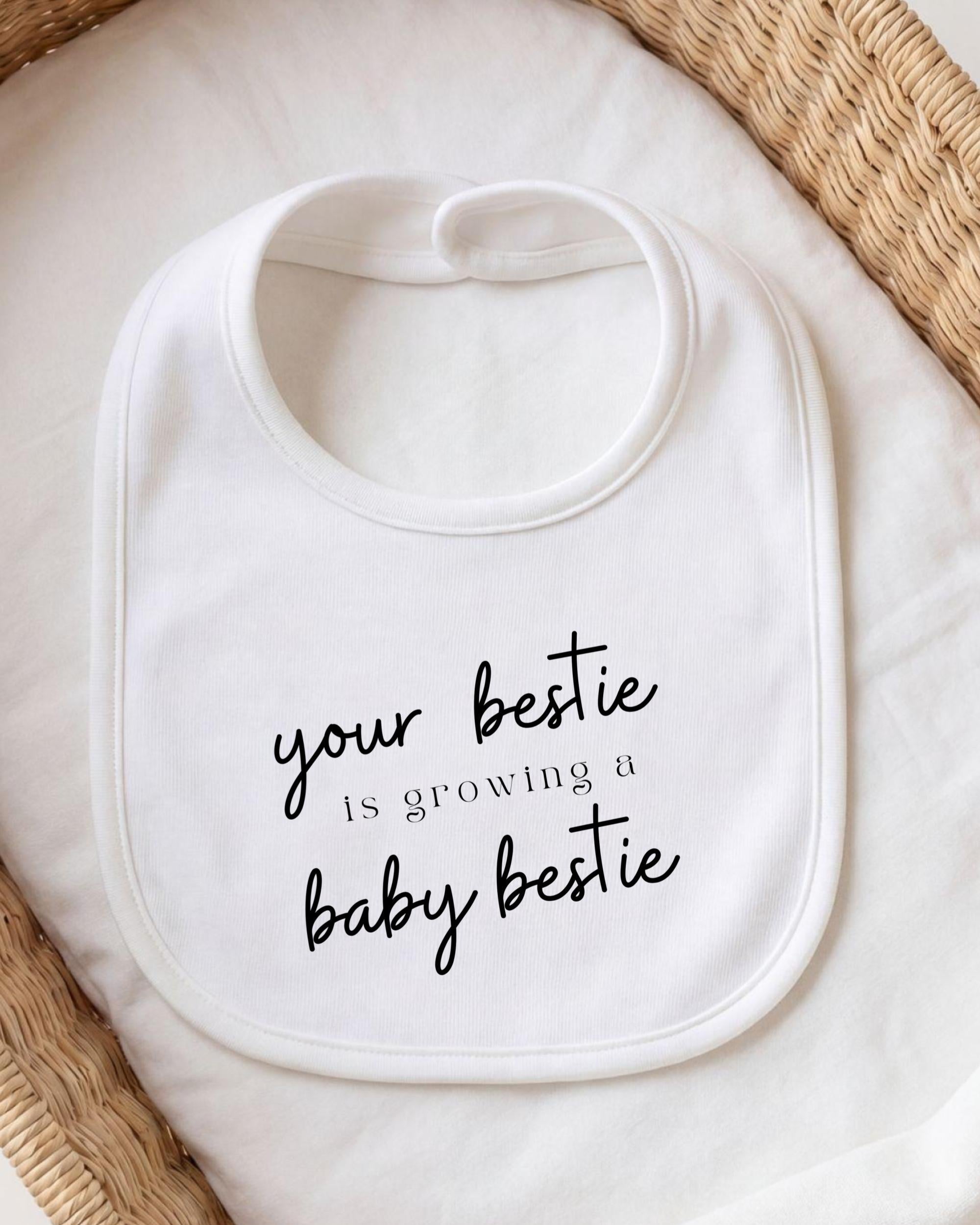 A white bib with the text "your bestie is growing a baby bestie" printed on it.