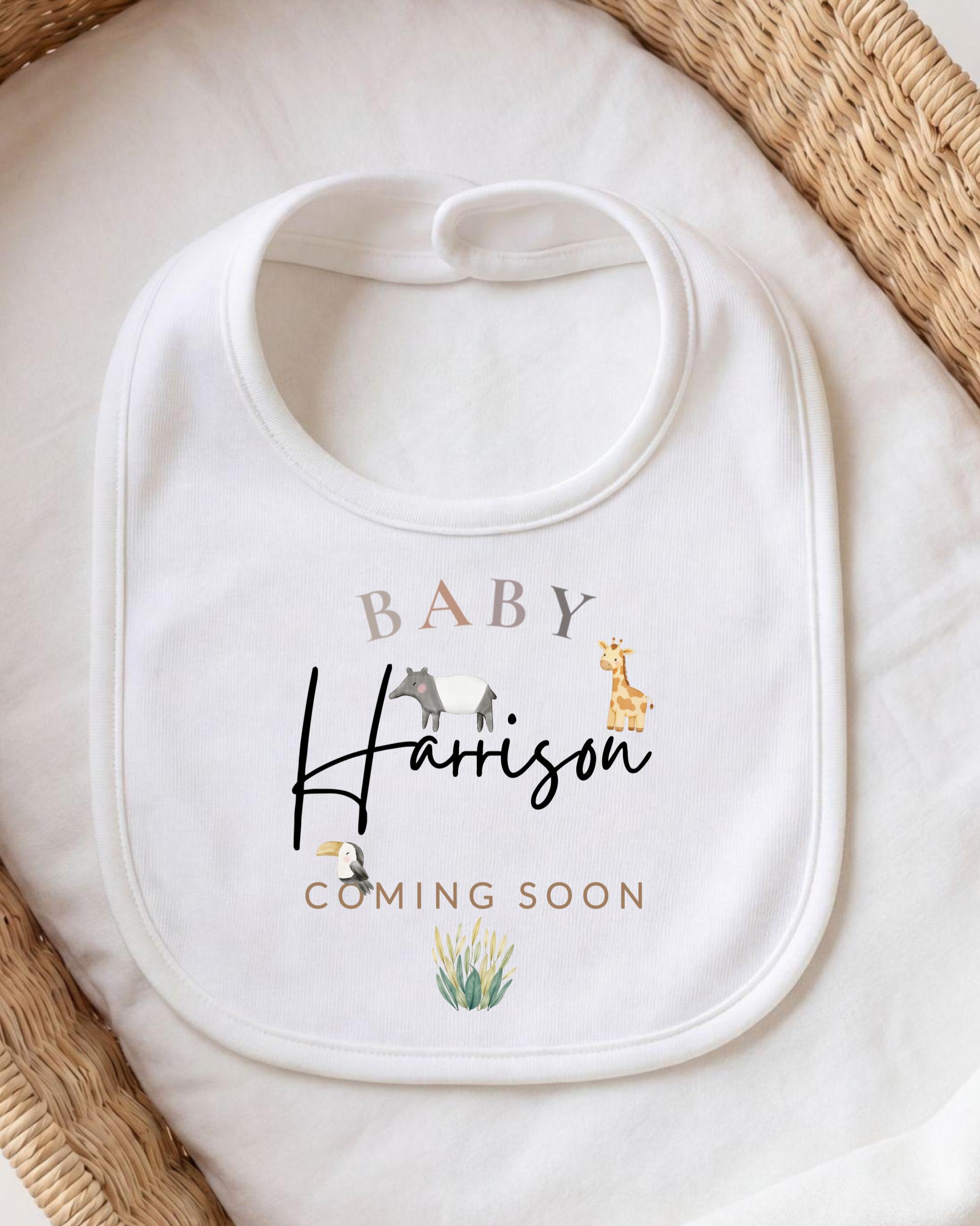 A white baby bib with the text "BABY Harrison COMING SOON" printed on it, along with illustrations of animals.