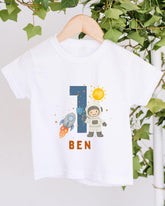 A white t-shirt with a graphic design featuring a space theme, including a rocket ship, a person in a spacesuit, and a number 1.