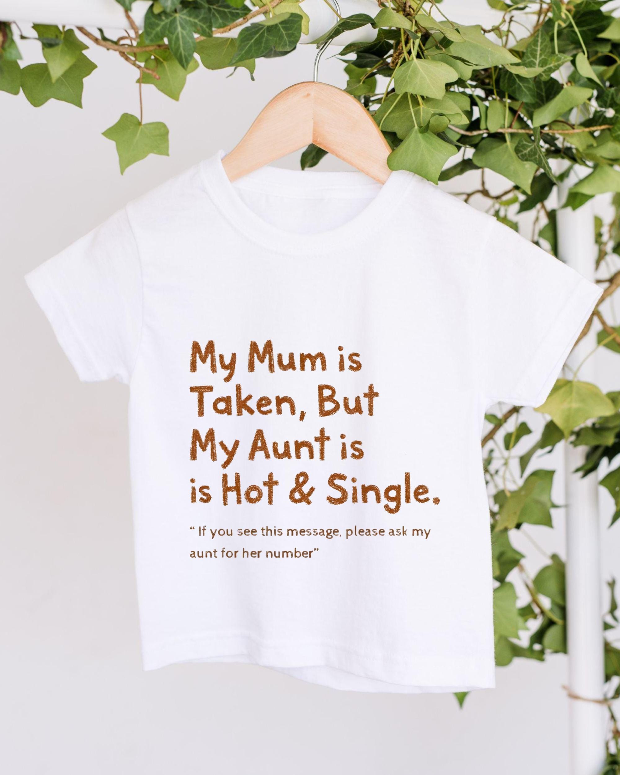 A white t-shirt with text printed on it, hanging on a wooden hanger. The text reads "My Mum is Taken, But My Aunt is Hot & Single. 'If you see this message, please ask my aunt for her number'".