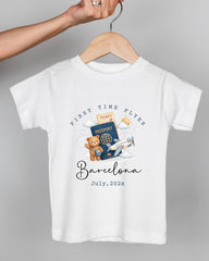 A white t-shirt with a graphic design depicting a teddy bear holding a passport and a plane, along with the text "First Time Flyer Barcelona July, 2023".
