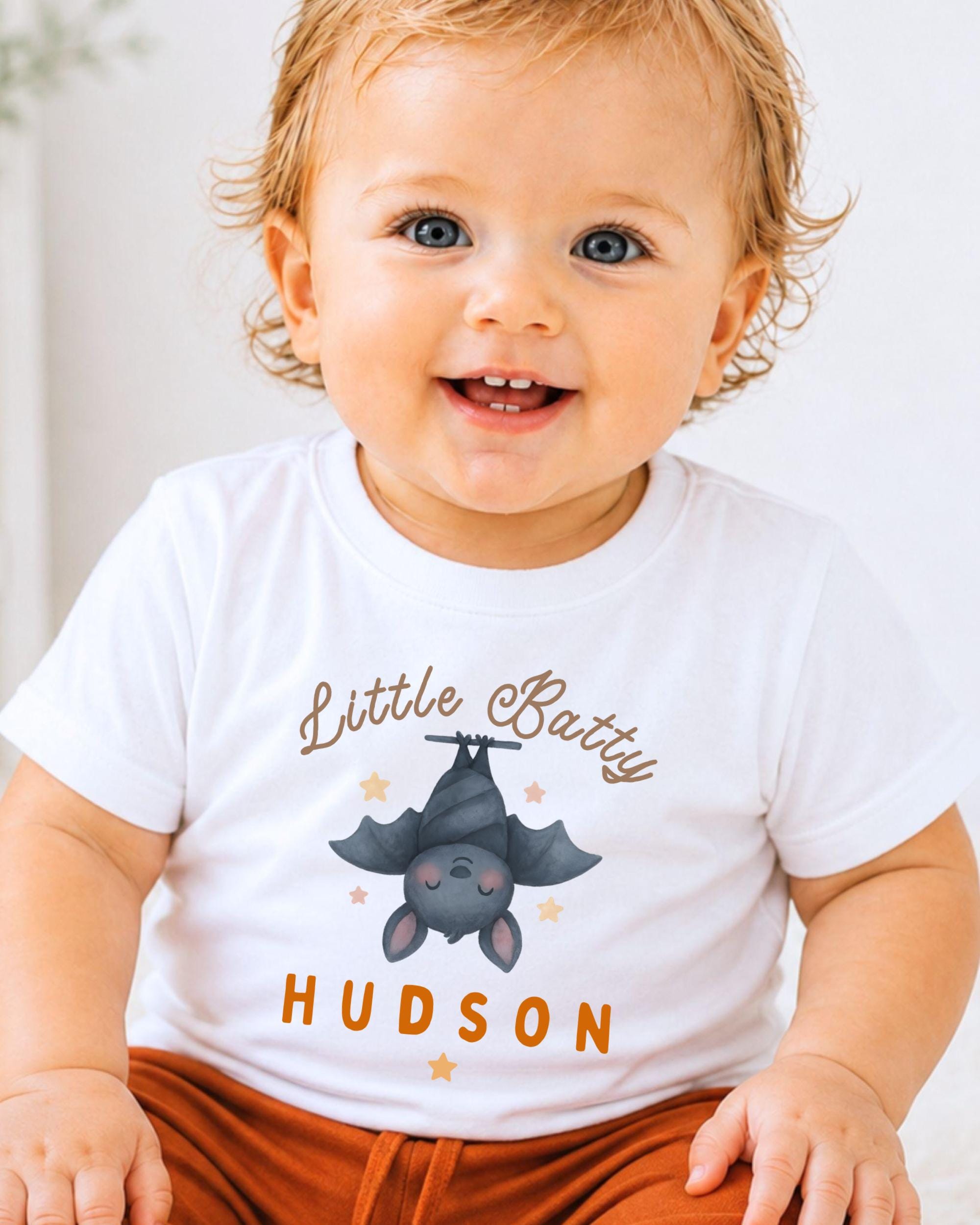 A smiling baby boy wearing a white t-shirt with a bat graphic and the name "Little Betty" printed on it.