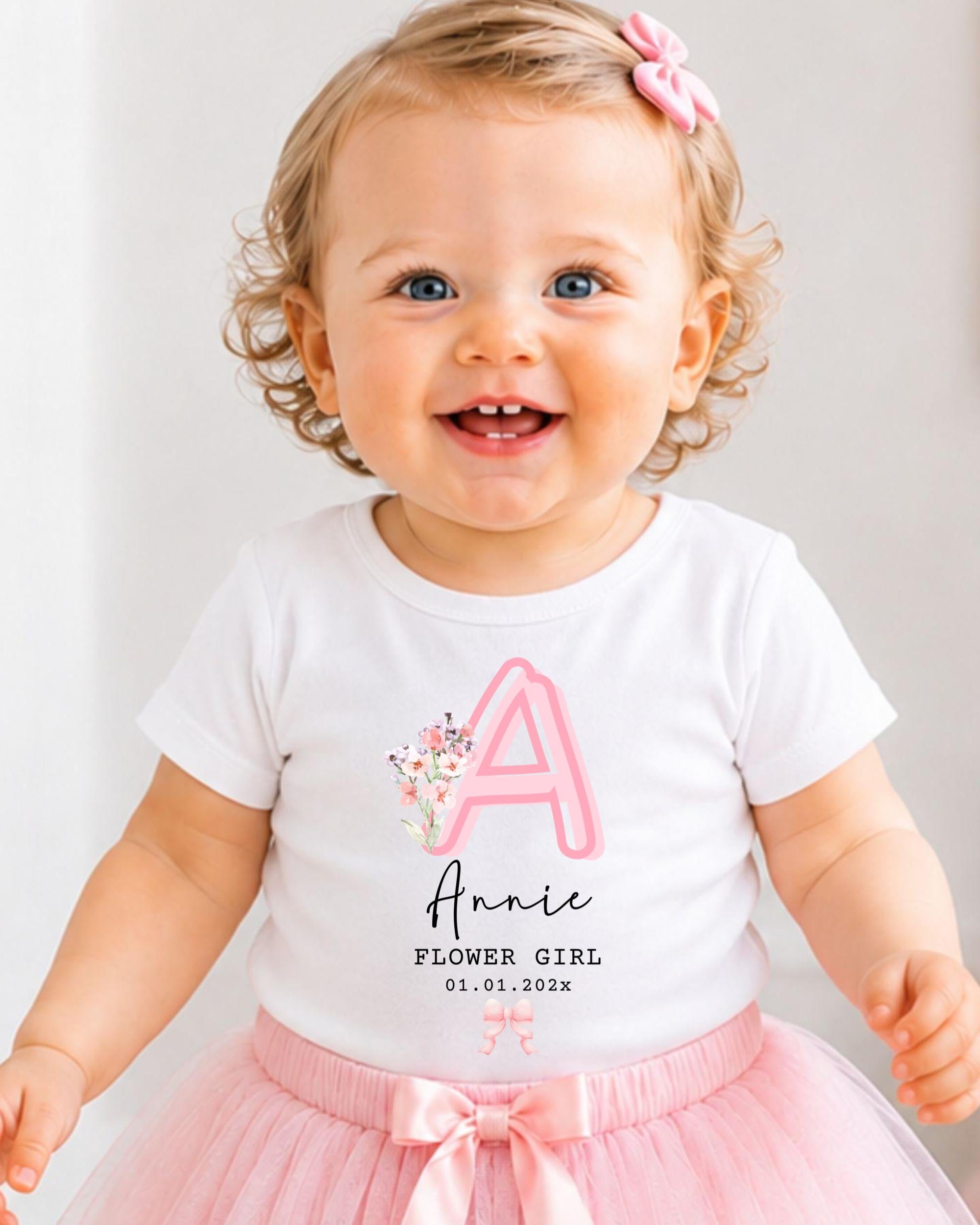 A smiling baby girl wearing a white t-shirt with a pink letter "A" and a pink tutu skirt, with a pink bow in her hair.